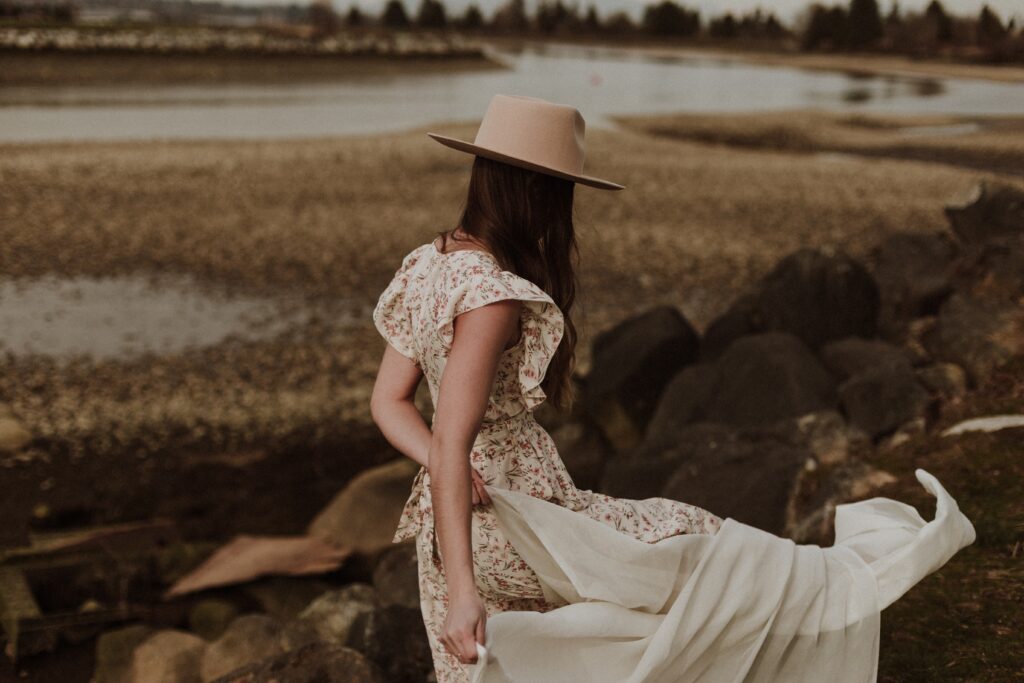 Woman wearing a floaty patterned white dress holding a white drape in the wind and wearing a cowboy hat at the beach