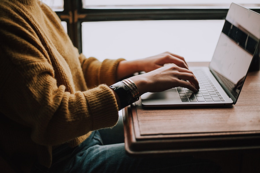 A view of a woman sitting at a wooden desk in front of a laptop, where she is doing her venue sourcing. She is wearing a knit pumpkin orange sweater, bangles, and teal pants.