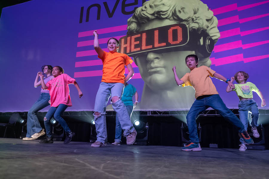 Six children wear blue jeans and brightly colored T-shirts as they perform a dance sequence on stage with a large LED screen behind them, featuring a large image of the head of Michelangelo's David sculpture wearing a VR headset with the word "HELLO" across the eyepiece. (Performed on stage at the CTCC)