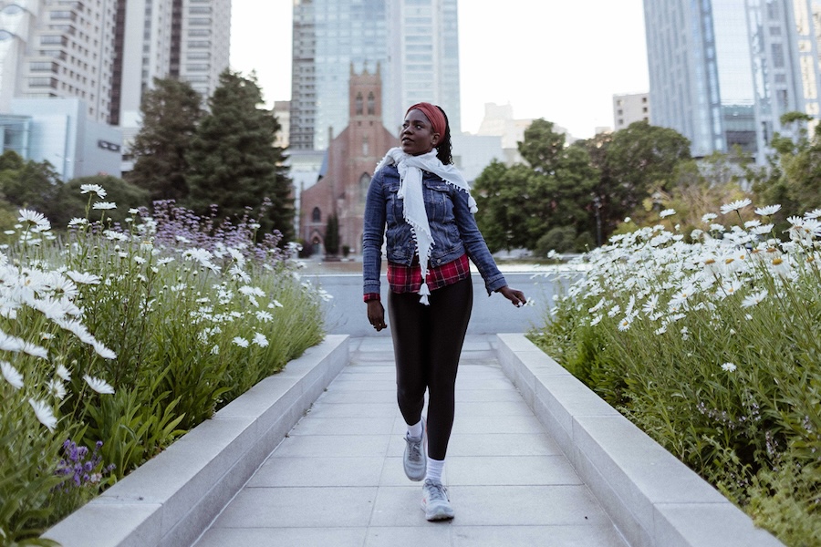 A woman is in the center and foreground of the photo, with large patches of daisy flowers on either side of her. In the middle ground, Saint Patrick's Church is visible with large trees on either side. Skyscrapers can be seen in the background. This kind of leisurely stroll through top city sites is a great bleisure addition to business events.
