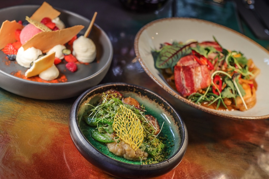 Three plates of vegan food, one showing a dessert with dollops of white and pink cream with strawberries, raspberries, and blueberries topped with a crispy pastry, and two others with leafy greens and roasted vegetables. Poland is known for its vegan cuisine.