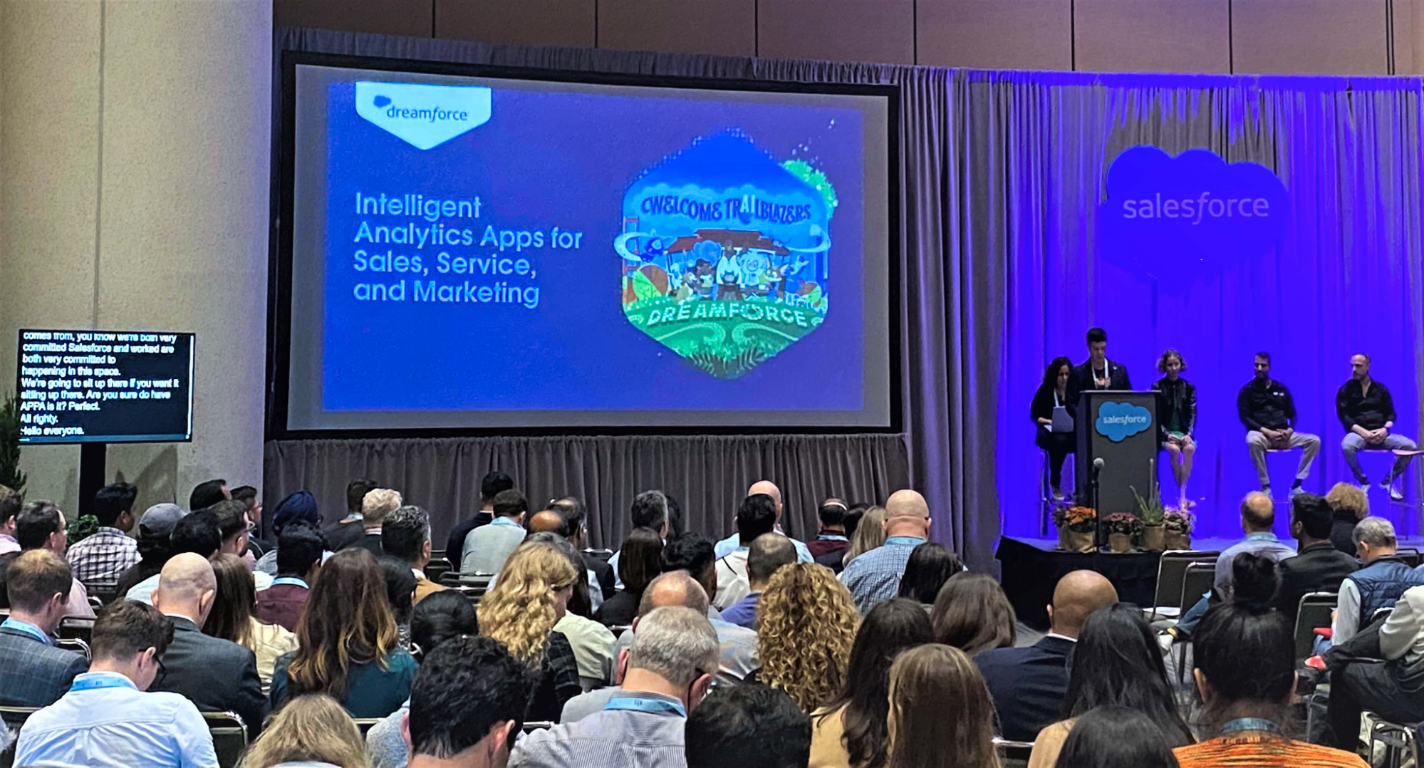 A view of a Dreamforce event from behind the audience, looking ahead at the stage at the far right, a large screen displaying a slideshow in the center, and a small monitor to the left displaying Wordly's live captioning.
