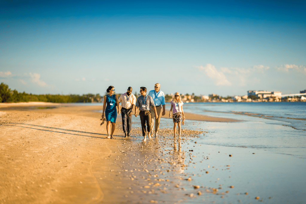 A group of five attendees takes a break from their coastal meeting to stroll down the beach without shoes.