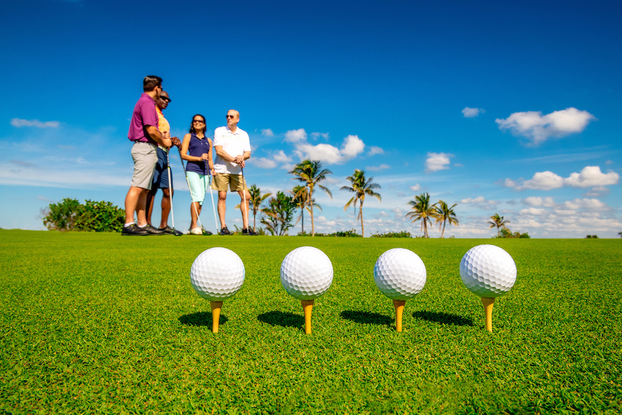Four golf balls are teed up in a line on the grass, with four golfers and multiple palm trees visible in the sun-drenched background.