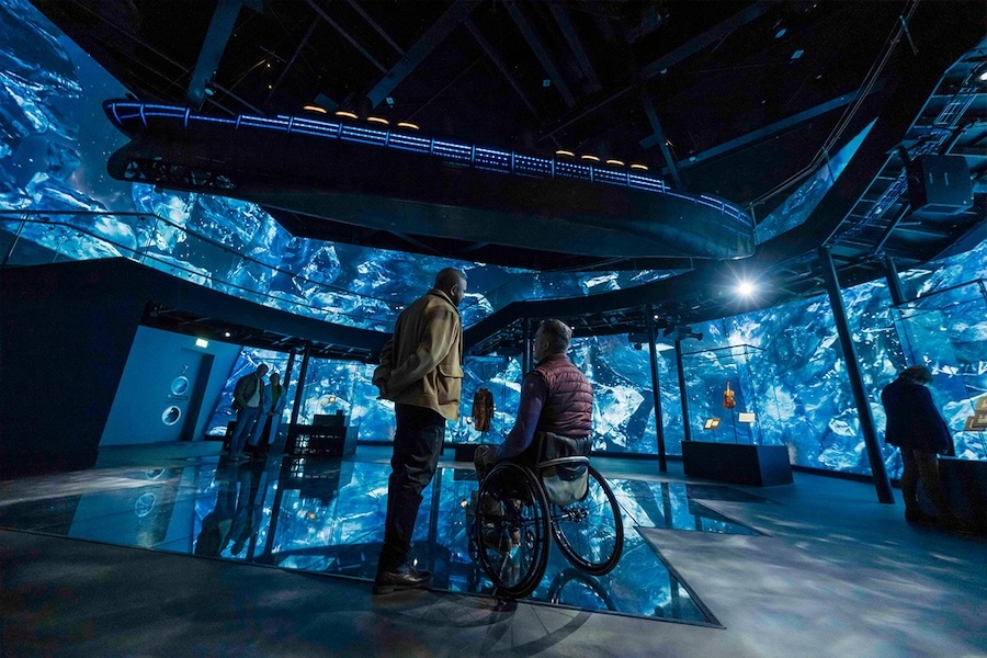 An interior room inside Northern Ireland's Titanic Belfast, featuring a large overheard recreation of the ship and projection imagery across the walls suggestive of blue water and ice.