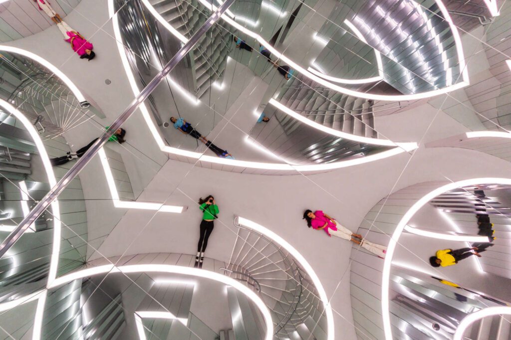 Gallery visitors lie on the floor looking up at their reflection on the ceiling, with curved patterns of geometric maze walls surrounding them. The walls are mirrored and do not reach the ceiling, in this way allowing their patterns to be viewed from the ceiling mirror.