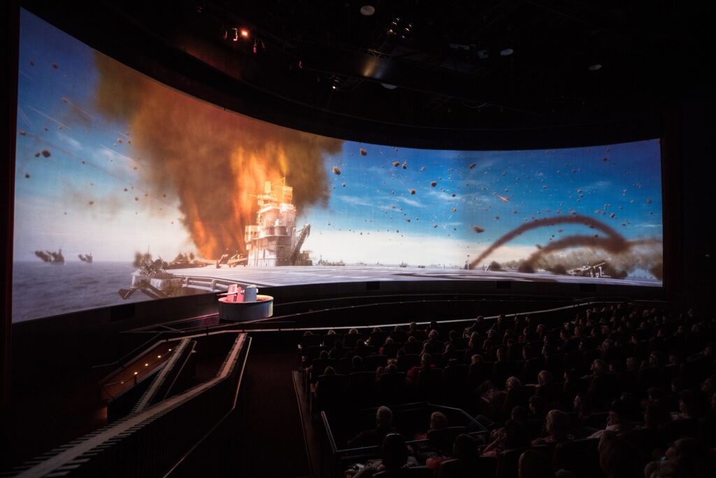 A large screen showcasing a wartime scene at sea, with darkened theatre seats barely visible in the foreground. The mechanized enhance the immersive experience.