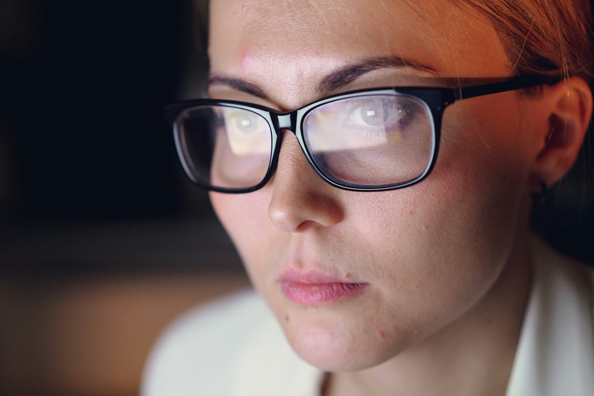 Close-up shot of a woman in professional attire wearing glasses using a computer in dark office room.